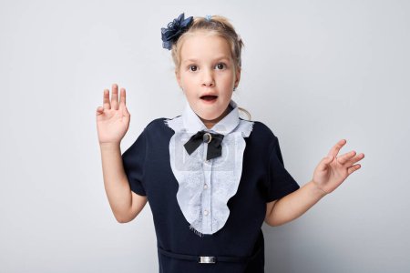 Téléchargez les photos : Petite fille surprise portant un uniforme scolaire, gesticulant les mains sur un fond clair. - en image libre de droit