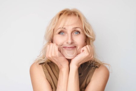 Téléchargez les photos : Portrait d'une femme blonde souriante avec les mains sur les joues, regardant rêveuse sur un fond blanc. - en image libre de droit