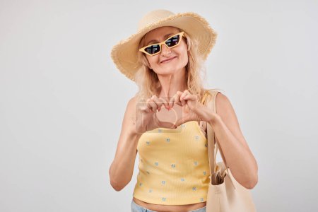 Foto de Mujer sonriente en sombrero de verano y gafas de sol haciendo forma de corazón con las manos en el fondo claro. - Imagen libre de derechos