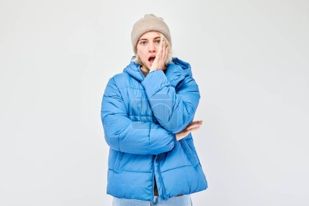 Téléchargez les photos : Jeune femme en tenue d'hiver regardant surpris avec la main sur la joue sur un fond blanc. - en image libre de droit