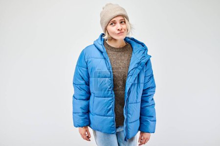 Young woman in blue puffer jacket and beanie posing with a curious expression on a plain background.