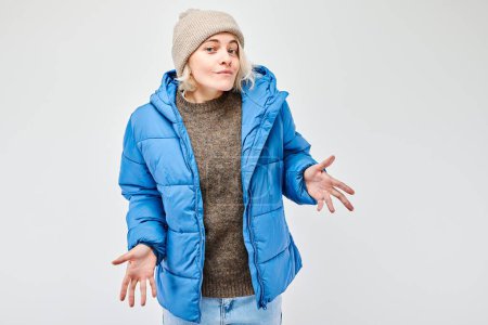 Woman in blue winter jacket and beanie shrugging with uncertain expression on a white background.