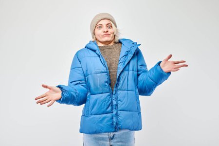 Femme en veste d'hiver bleue et bonnet haussant les épaules avec une expression incertaine sur un fond blanc.