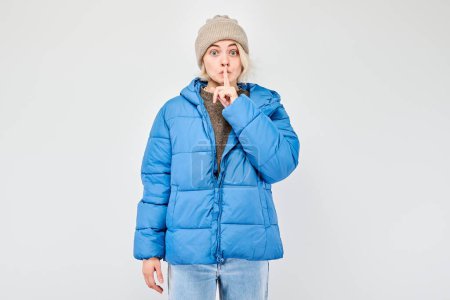 Woman in blue winter jacket gesturing silence with finger on lips, isolated on light background.