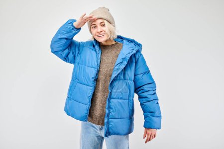 Persona sonriente en chaqueta de invierno azul mirando hacia otro lado con la mano en la frente contra un fondo claro.