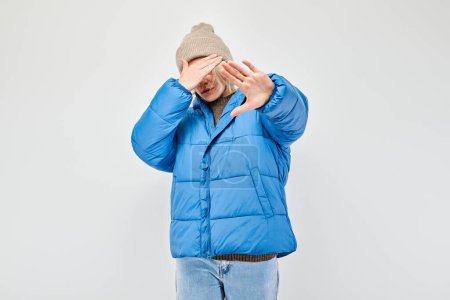 Man in blue jacket and beanie covering face with hands, white background.
