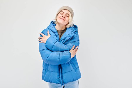 Téléchargez les photos : Femme en veste d'hiver bleue et bonnet se serrant dans ses bras et souriant sur un fond clair. - en image libre de droit