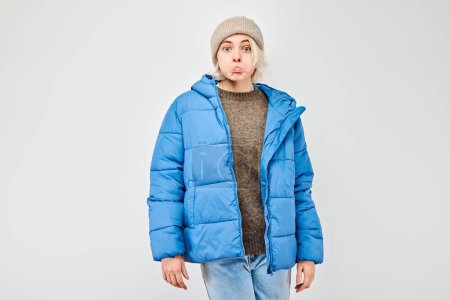 Téléchargez les photos : Jeune adulte dans une veste gonflante bleue et bonnet fait une grimace drôle sur un fond blanc. - en image libre de droit