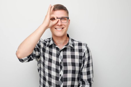 Téléchargez les photos : Homme joyeux en chemise à carreaux faisant OK geste sur l'œil, souriant à la caméra sur fond blanc - en image libre de droit