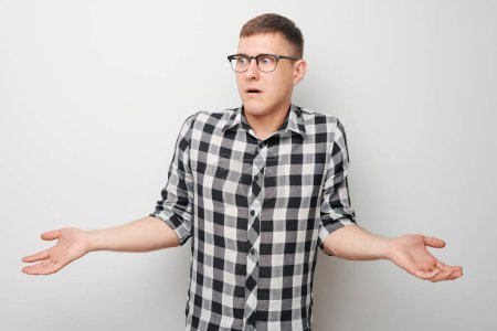 Photo for Confused young man in checkered shirt shrugging shoulders on white background expressing uncertainty - Royalty Free Image
