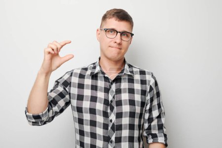Téléchargez les photos : Homme en chemise à carreaux faisant un geste de petite taille avec les doigts, fond neutre. - en image libre de droit