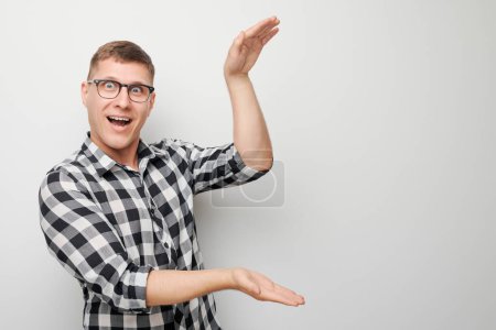 Téléchargez les photos : Joyeux jeune homme en lunettes et chemise à carreaux geste avec les mains sur un fond uni. - en image libre de droit