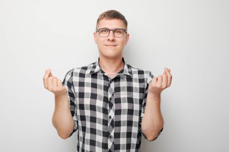 Téléchargez les photos : Jeune homme en chemise à carreaux et lunettes croisant les doigts pour bonne chance sur un fond blanc. - en image libre de droit