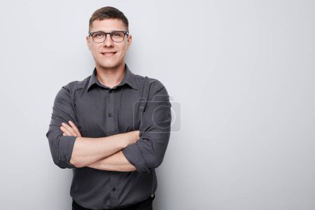 Téléchargez les photos : Jeune homme confiant dans des lunettes et une chemise grise, bras croisés, debout sur un fond uni. - en image libre de droit