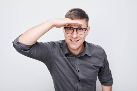 Photo for Smiling young man in glasses looking into the distance with hand above eyes on a plain background. - Royalty Free Image