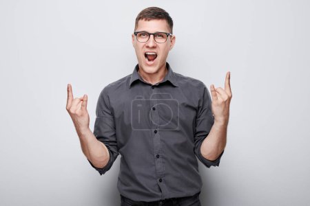Téléchargez les photos : Jeune homme excité dans des lunettes gesticulant pierre sur signe sur un fond gris. - en image libre de droit