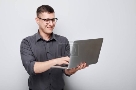 Photo for Professional man in glasses using laptop against a gray background with a focused expression. - Royalty Free Image