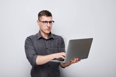 Téléchargez les photos : Homme professionnel dans des lunettes en utilisant un ordinateur portable sur un fond gris avec une expression ciblée. - en image libre de droit