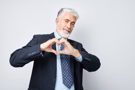 Photo for Mature businessman making heart shape with hands, smiling, on grey background. - Royalty Free Image