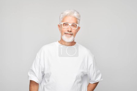Téléchargez les photos : Homme âgé avec des lunettes portant un t-shirt blanc, souriant sur un fond gris. - en image libre de droit