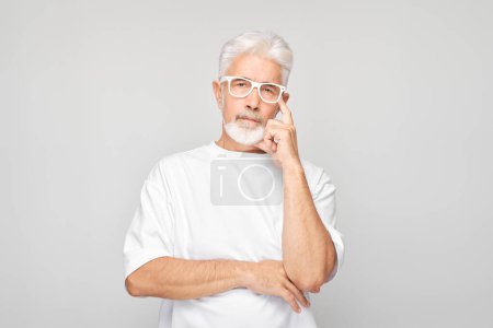 Téléchargez les photos : Homme âgé confiant avec des lunettes sur fond gris regardant la caméra avec une expression réfléchie - en image libre de droit