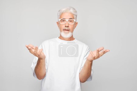 Téléchargez les photos : Homme âgé aux cheveux gris en lunettes et chemise blanche faisant un geste incertain avec les mains levées - en image libre de droit