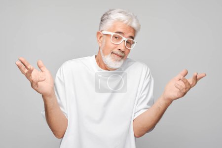 Téléchargez les photos : Homme âgé confus avec des lunettes haussant les épaules sur un fond gris. - en image libre de droit