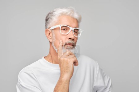 Téléchargez les photos : Homme âgé pensif avec des cheveux blancs et des lunettes, main sur le menton, isolé sur fond gris. - en image libre de droit