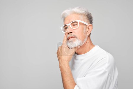 Foto de Hombre mayor pensativo con pelo blanco y gafas, mano sobre barbilla, aislado sobre fondo gris. - Imagen libre de derechos
