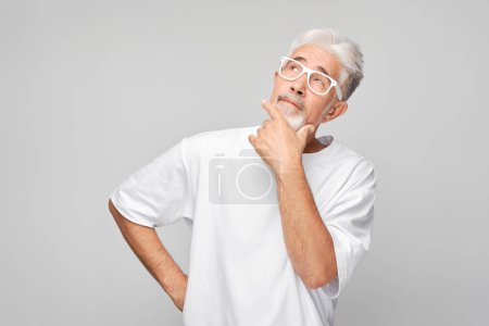 Photo for Elderly man in white shirt and glasses looking up thoughtfully, hand on chin, on gray background. - Royalty Free Image