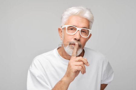 Foto de Hombre mayor con pelo blanco y gafas haciendo gesto de silencio sobre fondo gris. - Imagen libre de derechos