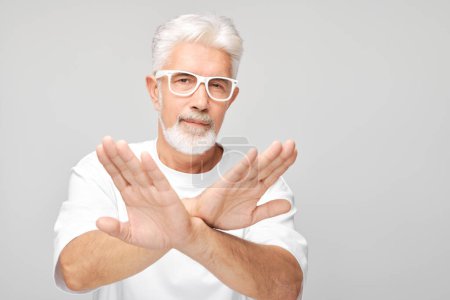 Selbstbewusster Senior mit Brille macht Stop-Geste mit verschränkten Armen auf grauem Hintergrund