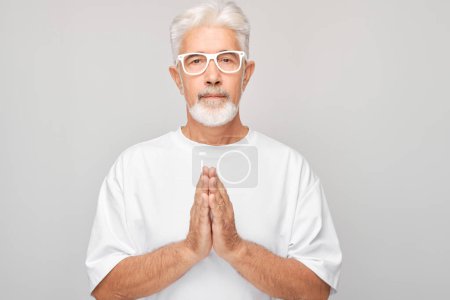 Téléchargez les photos : Homme âgé en chemise blanche et lunettes avec les mains en position de prière sur un fond gris. - en image libre de droit