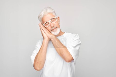 Photo for Senior man with glasses pretending to sleep, standing against a gray background. - Royalty Free Image
