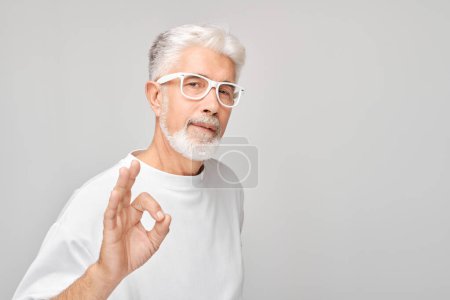 Foto de Hombre mayor con gafas haciendo gesto OK sobre un fondo gris. - Imagen libre de derechos
