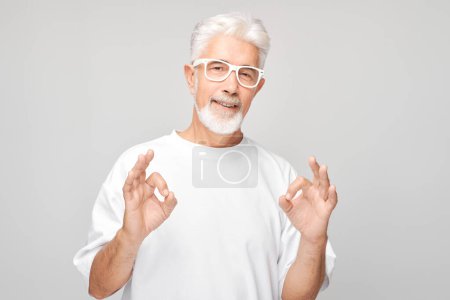 Téléchargez les photos : Senior homme avec des lunettes faire un geste OK sur un fond gris. - en image libre de droit