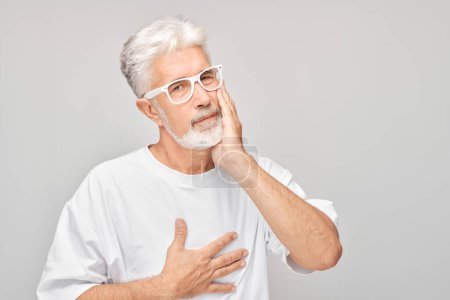 Téléchargez les photos : Homme âgé avec des lunettes touchant la joue dans l'inconfort, douleur dentaire, sur un fond gris. - en image libre de droit