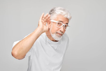 Téléchargez les photos : Homme âgé avec des lunettes tasses main derrière l'oreille pour mieux entendre, sur un fond gris. - en image libre de droit