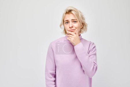 Téléchargez les photos : Jeune femme pensive dans un pull violet, la main sur le menton, regardant réfléchi sur un fond clair. - en image libre de droit