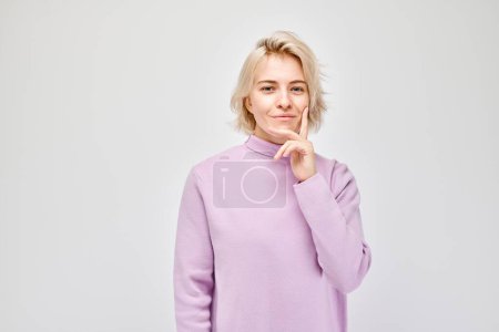 Photo for Pensive young woman in a purple sweater, hand on chin, looking thoughtful on a light background. - Royalty Free Image