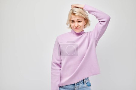 Foto de Mujer joven confundida arañándose la cabeza, llevando un cuello alto rosa sobre un fondo claro. - Imagen libre de derechos