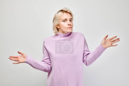 Foto de Mujer joven confundida en un cuello alto púrpura encogiéndose de hombros con las manos arriba, aislada sobre un fondo blanco - Imagen libre de derechos