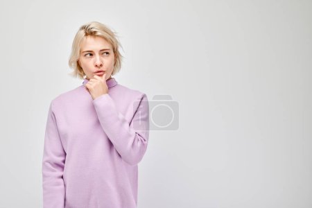 Téléchargez les photos : Jeune femme réfléchie en pull violet avec un doigt sur le menton, isolée sur un fond clair. - en image libre de droit