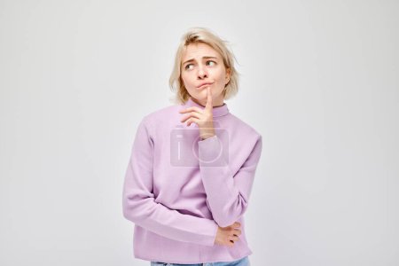 Téléchargez les photos : Jeune femme réfléchie en pull violet avec un doigt sur le menton, isolée sur un fond clair. - en image libre de droit