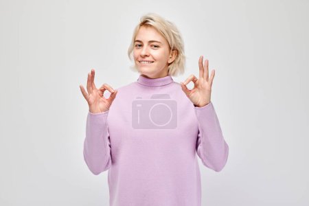 Téléchargez les photos : Portrait d'une femme blonde réjouissante montrant un geste OK avec ses doigts sur un fond blanc - en image libre de droit