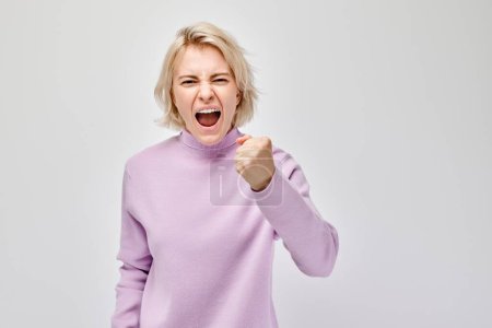 Foto de Mujer enojada en un suéter púrpura apretando los puños y gritando, aislada sobre un fondo blanco. - Imagen libre de derechos