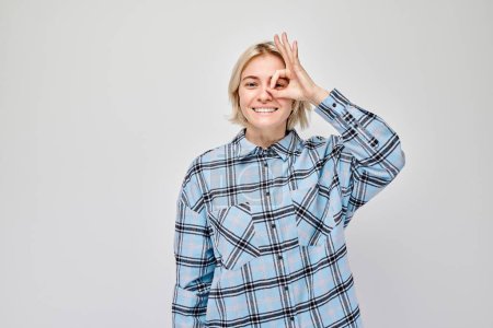 Foto de Mujer sonriente haciendo gesto OK sobre el ojo, usando camisa a cuadros sobre fondo blanco. - Imagen libre de derechos