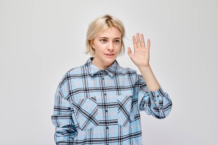 Téléchargez les photos : Femme blonde en chemise à carreaux gesticulant bonjour avec sa main, isolée sur un fond clair. - en image libre de droit