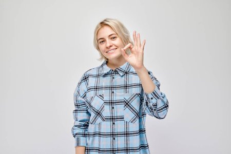 Foto de Mujer sonriente en camisa a cuadros haciendo signo OK con la mano sobre un fondo claro. - Imagen libre de derechos