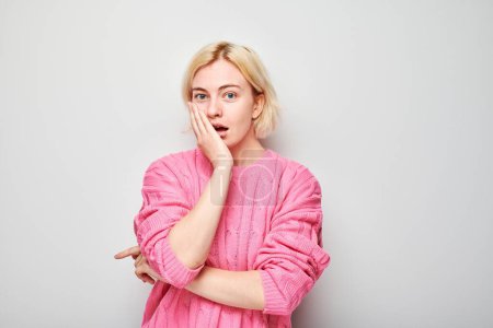 Surprised young woman in pink blouse with hand on cheek, white background.